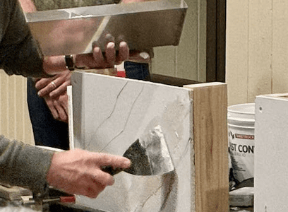 A close up of two hands, one holding a metal tray and the other uses a putty knife to apply plaster to a small model of a wall