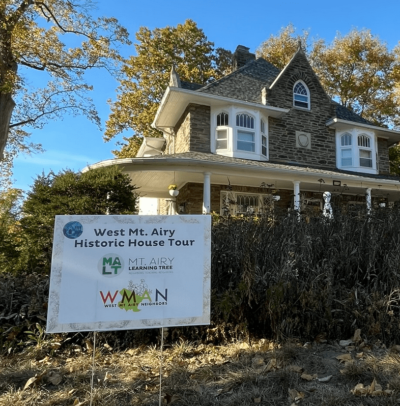 A lawn sign for the West Mt Airy Historic House Tour is placed in the foreground in from of some green bushes. Behind the bushes a stone house with a covered porch and white trim can be seen.